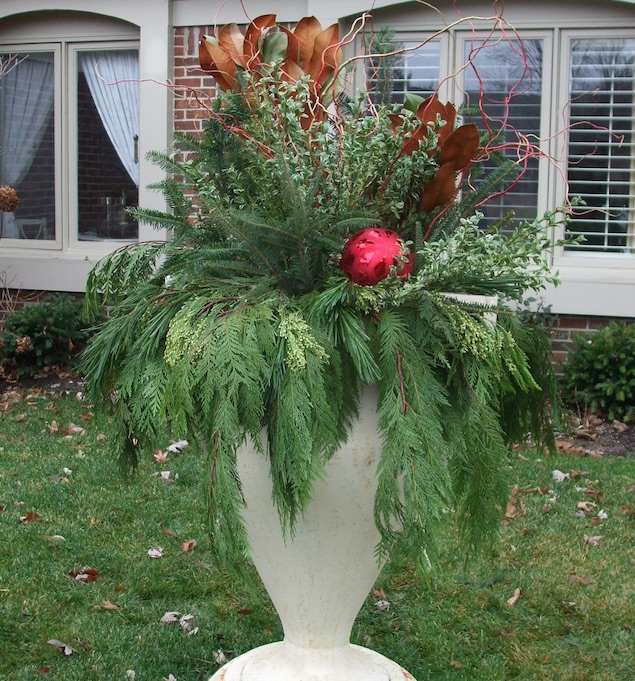 A dramatic look for an antique urn comes from select evergreen boughs, magnolia branches, color stems and an outdoor ornament. (C) Jo Ellen Meyers Sharp